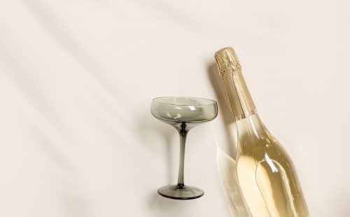 Champagne glasses tinted grey glass, Champagne or sparkling wine bottle on beige background. Festive drink concept, glare and sunlight. Modern wine colored glasses. Creative flat lay, copy space
